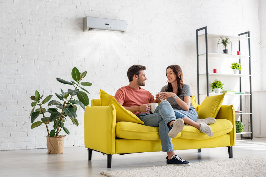 happy young couple talking and holding cups while sitting on yellow sofa under air conditioner at home